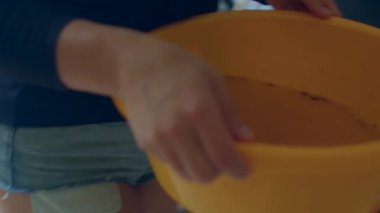 Yatak odasında sarı leğen tutan beyaz kadın, güneşli dairede çamaşırları hazırlayan plastik küveti tutan ellerin yakın çekimi, sıradan kot şortlar, rahat pratik ruh hali, yavaş yerli koreografi