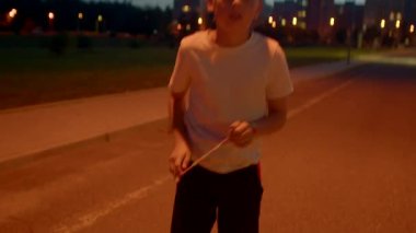 Turuncu sokak lambasının altında boş bir yolda duran beyaz bir genç, elleri ceplerinde, yalnız gece havasında.,