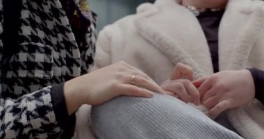 Müstakbel beyaz gelin ve Asyalı arkadaş aynı bankta oturuyor neşeli gülüşler ve rahatlatıcı el dokunuşları, yumuşak kürk manto ve houndstooth ceket kontrastı, ellere yakın çekim ve gülümseme, şehir meydanı titreşimi