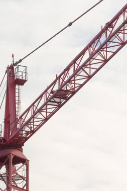 Gökyüzüne karşı kırmızı inşaat vinci, çerçevede çelik kafes patlaması ve operatör taksisi, sessiz bulutlu ışık, örülmüş donanım ve görünür çekiş, arka planda kentsel yapı iskeleti, atmosfer