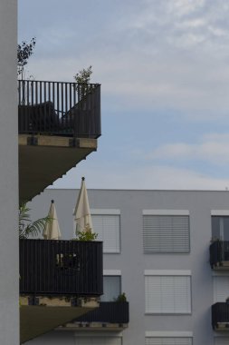 Bitkilerin ve şemsiyelerin olduğu balkon köşesi şehir cephesi ve gökyüzü, metal korkuluklara konmuş konforlu yeşillik, yumuşak bulut yansımaları, ince gölgeler ve rahat şehir terası öneren sabah ışığı