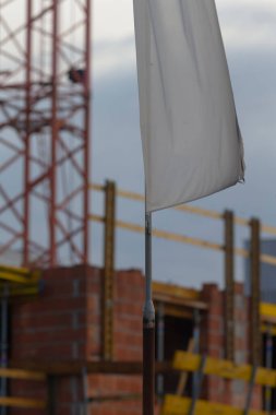 Bölgede dalgalanan beyaz güvenlik bayrağı, ön planda kapalı kumaş detayı, arka planda bulanık tuğla duvar ve yapı iskelesi, kereste biçiminde ve çevresel direkler görünür, esinti hareketi ve sessiz gökyüzü