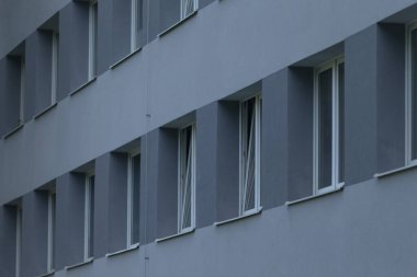 Gri cephedeki uzun pencereler ritmik yineleme ve doğrusal kompozisyon yaratıyor, geniş cephe görünümü modülleri vurguluyor ve düzensel mimari veya örüntü odaklı tutarlı aralıklar oluşturuyor