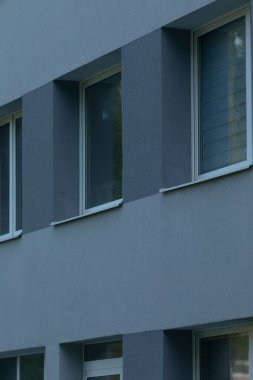 Gri bina cephesi, kapalı pencereler, dikey koyları gösteren temiz modern dış görünüş ve yumuşak bulutlu ışık altında yansıtıcı camlar, minimal çizgilerin mimari çalışması ve sessiz palet