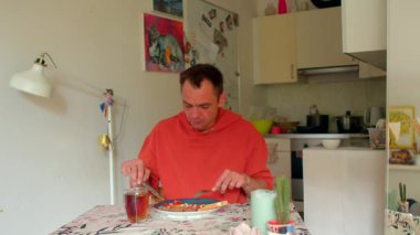 Beyaz adam mutfak masasında kahvaltı yapıyor. Serbest yazar, çay yudumlarken notlar alıyor. Kırmızı süveter, rahat.