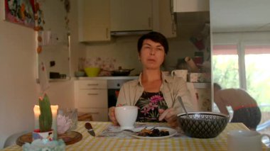 Beyaz kadın mutfak masasında otururken pencereden dışarı bakıyor, elinde fincanla beyaz bir adam gibi kapıya bakıyor.