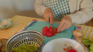 Beyaz kadın mavi tahtada olgun domatesleri doğradı, desenli kaseyi doğranmış meyvelerle doldurdu, kareli önlük giydi.,