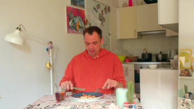 Mutfak masasında beyaz bir adam yemek yerken içecekten örnekler alıyor. Yemek tarifi test eden kişi tuhaf bir ifadeyle lezzeti değerlendiriyor.