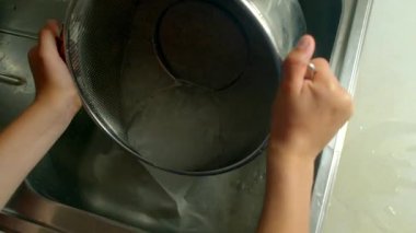 Eller tencere ve kevgir suyunu lavaboya boşaltmak için eğiliyor, metal yüzeyi inceliyor, mutfak temizliği yapıyor.