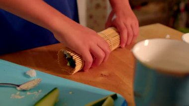 Beyaz çocuk suşiyi sımsıkı sarmak için bambu hasırı, salatalık dilimleri ve düzgün eller kullanıyor.