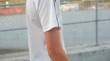 Zincirli tel örgülü, beyaz, çitten geçen genç. Bileği bandajlı, sıradan tişörtlü, banliyö kaldırımlı.