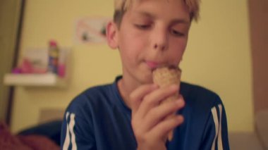 Kafkasyalı çocuk yatak odasında dondurma külahı yalıyor, küçük külah sıkı tutulmuş, yavaş tadını çıkarıyor, sıcak tonlar, rahat gençlik havası.