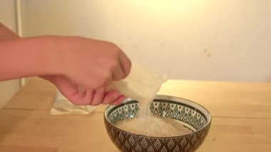 Yakın çekim eller pirinç torbasını desenli kaseye boşaltıyor, sıcak duvar ışığı, hassas mutfak hareketi ve düzenli.