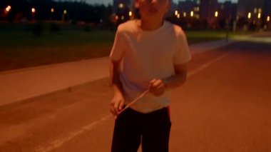 Genç çocuk, turuncu parıltının altında boş bir sokakta duruyor, elleri ceplerinde ve rahat duruş, uzak şehir ışıkları.