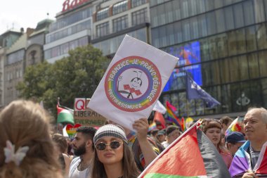 Prag eylemcisinin renkli gurur yürüyüşü elinde amblem işareti, parlak kıyafetler ve güneş gözlüğü takan kalabalık, bayrak ve afişler sallıyor, gülümseyen CZ, Prag, Na prikope, 2.8.25