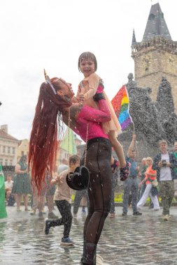 Omuzlarında tek boynuzlu at maskesi, gökkuşağı bayrağı, çeşme spreyi, tezahürat yapan kalabalık, ıslak kaldırım taşı renkli kostümler ve gotik kule CZ, Prag, Na prikope, 2.8.25