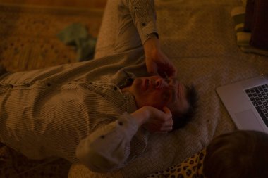 Adam yerde yatıyor, dizüstü bilgisayar açık, alnında kehribar lamba parıltısı, altında desenli halı yumuşak ışık saçan dizüstü bilgisayar, yorgunluk ve iç gözlem gösteren ifade yüzlü, gecikme