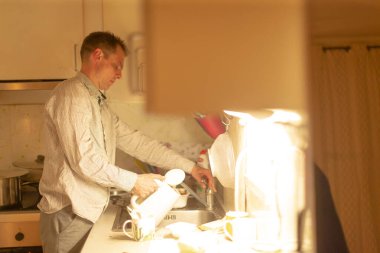 Adam gece bulaşık makinesini boşaltıyor, tabakları ve bardakları dolaba koyuyor, sıcak mutfak ışığı altında düzenli hareketler düzenliyor, ev düzenini sıfırlıyor ve etkin bir şekilde temizlik yapıyor, dikkatli bir şekilde ev içi temizlik yapıyor.