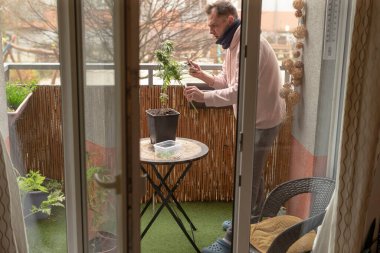 Beyaz adam balkon bitkilerini enerjik bir şekilde tartışıyor, tencere ve küçük masaya el kol hareketi yapıyor, ifade edici duruş, görünmeyen komşuyla konuşuyor, şehir manzarası, günlük süveter, sosyal yardım havası.,