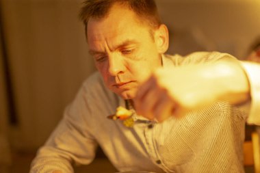 Beyaz adam tek başına çorba içiyor, emekli öğretmen kehribar lambanın altında ahşap masada oturuyor düşünceli bir ifadeyle kaşığı karıştırıyor, ılık bir mutfak akşamı yumuşak ışıklandırmalı, derin derin düşünüyor.