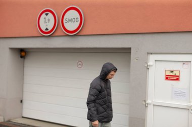 teenager walking past garage door under cloudy sky, wearing hooded jacket, hands in pockets, muted facade with circular signs, empty sidewalk, residential block in background, introspective posture