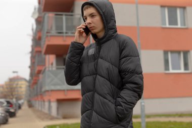 Dışarıda telefonla konuşan genç bir çocuk, apartman zemininde kapüşonlu bir kabarık ceket, derin düşünceli bir ifadeyle kaldırımda duruyor, soğuk bulutlu bir atmosfer, gizli sokak resimlerini araştırıyor.
