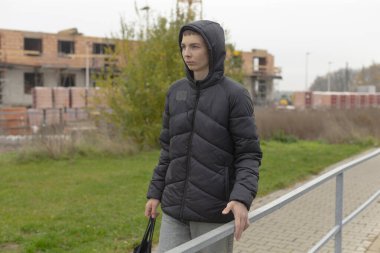 Genç bir çocuk, elinde sırt çantası ve inşaat sahasının yanındaki not defterine yaslanıyor. Kapüşonlu bir ceket ve düşünceli bir bakış. İlerleme, sonbahar yeşilliği ve bulutlu gökyüzü, yansıtıcı hava.