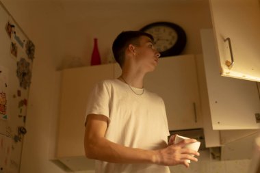 Tezgaha uzanan genç beyaz erkek, saat ve dolaba doğru bakıyor, sıcak mutfak ışığı altında düşünceli duruş, geç akşam ritüeli, küçük dairede yumuşak gölgeler ve durgunluk.,