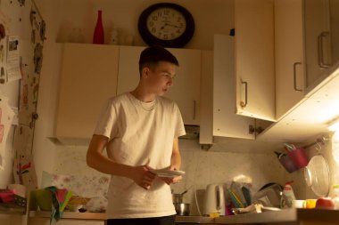 Gençler mutfak tezgahında yemek pişiriyor ve tarifi takip ediyor. Acemi şef enerjisi, çatal bıçaklar ve dağınık malzemeler, parlak ışık altında odaklanmış duruş, saat ve kiler görünür.,