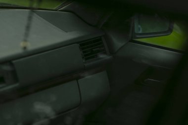 Boş yolcu koltuğu ve renkli pencereden görülebilen gösterge paneli, yapraklı şehir ortamında dinlenme aracında müfettiş rolü, sessiz yeşil yansımalar ve desenli döşeme detayları