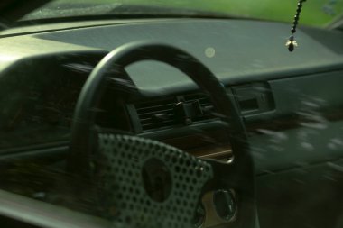 Torpido gözünde tespih asılı direksiyon, taksi şoförü ritüeli ima edilmiş, sessiz koruyucu tılsım, dokulu direksiyon kirişi ve park yansımalı yansıtıcı yan ayna.