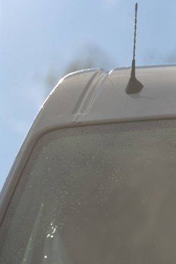 Şafak vakti ıslak minibüsün ön camı anten ve dağınık yağmur damlalarını gösteriyor, yumuşak mavi gökyüzü yansıması, cam üzerindeki ince kirlilik, soğuk ışık ve metal çatı detaylarıyla şehir sabah banliyösü atmosferi.