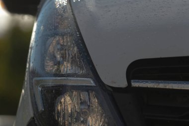 Aracın ön tarafındaki ıslak far krom kaplama, lens dokusu ve küçük damlacıklar, karamsar sığ derinlik ve öğleden sonra ışığı, endüstriyel yol kenarı ayarı öneriliyor.