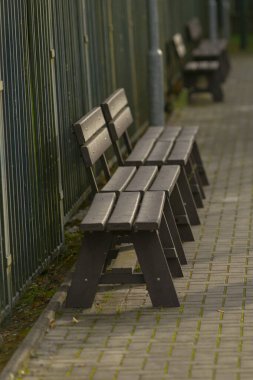 Boş ahşap park bankları asfalt yol kenarındaki metal çitler boyunca hizalanmış ıslak kaldırım taşları, yumuşak sabah ışığı, sessiz yeşil arka plan, sığ alan derinliği, sakin kompozisyon, kentsel yalnızlık