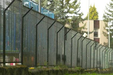 Beton zemin üzerinde açılı çivileri olan uzun metal çitler, zincirli paneller ve eğimli güvenlik çubukları güçlü sınırlar yaratıyor. Arka planda görülen bulanık apartman bloğu, cesur kentsel