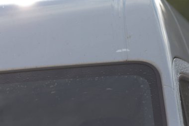 Kavisli metal, küçük su lekeleri ve parlak yansımaları gösteren güneşli minibüs çatısı kapanışı, sıcak açılı ışık şekli ve yüzeysel sonu vurguluyor, editoryal otomotiv detaylı çekimi.