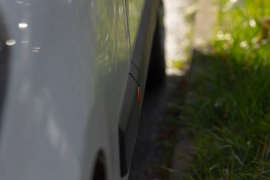 Çimenlerin yanında beyaz araba tekerleği, vücut panelinde görünür kehribar yansıtıcı, kaldırım ve asfalt yakın mahsul, banliyö yol kenarı detayı güneş ışığı ve gölge, otomotiv için uygun