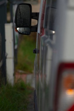 Kapalı kamyonet yan aynası gece karanlığında bulanık arka lambaları ve yol kenarındaki çimenleri gösteriyor. Yansıtıcı camlar trafiği yakalıyor, sığ alan derinliği, tozlu boya işi ve çamurluk, kentsel son mil çizikleri.