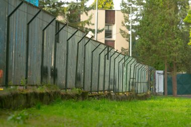 Çimenli çit boyunca çitler, açılı metal paneller ve örgüler, paslı üsler ve yeşil kapı mesafesi, yeşil alanı inşa edilmiş çevreden ayıran park sınırı, koruma ve koruyucu ruh hali,
