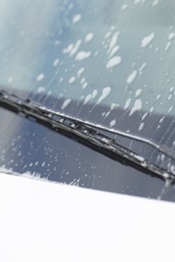 Sabun köpüklü ön cam temizleyici köpük, çizgi desenleri ve damlacıklar, berraklık onarım odağı, cam yüzeye yakın çekim ve bıçak etkileşimi, gündüz araba bakımı sahnesi