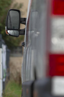 Islak yan ayna ve minibüs paneli yakın plan, cam damlacıklar ve çevrenin ince yansıması, boyalı yüzeyde görülebilen temas olmayan durulama dokusu, ayrıntılı hazırlık ima edildi, yumuşak arkaplan bokeh