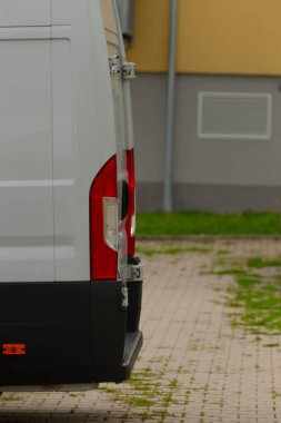 Arkadaki minibüs park yerini kapatın. Araç ve bina arasında dar bir boşluk, yakın zamanda yıkanan tuğla kaldırımlar, sarı cephe ve metal direkler, görsel öneri.