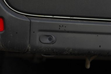 Algılayıcı kapaklı kirli arka tampon, plastik çerçevenin etrafında su izleri ve çamur birikintileri, boyalı yüzeye karşı görülen park sensörü, güvenlik ve estetik için denetim ve temizlik bağlamında,