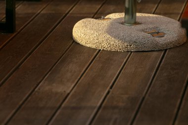 Güvertede güneşli teras şemsiyesi, metal çubuk tutan beton halka, sıcak altın ışık parke üzerinde uzun yumuşak gölgeler bırakıyor, boş kafe köşesi sakin bir sabah atmosferini çağrıştırıyor.