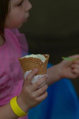 Profil çocuğunun naneli dondurmanın tadını çıkarması, peçeteye sarılı waffle külahının sıkıca kavraması, pembe gömlek ve sarı bileklik görünür, bulanık park arka planı, sıcak güneş ışığı ve sessiz odaklanma, samimi bir portre.