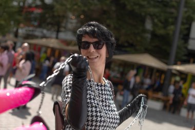 Sokak şarkıcısı mikrofonla şarkı söylüyor, av elbisesi giyiyor, siyah eldivenler takıyor ve güneş gözlüğü takıyor, kalabalık şehir meydanında geçen kalabalığa gülümsüyor, klasik müzik, karizmatik nişan, hayat dolu.