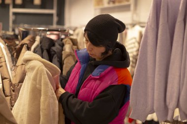 Müşteri ceketleri inceler, alışverişçiler mağaza ortamında zıt ceket tasarımlarını değerlendirir, alıcı mağazanın etrafını incelerken farklı ceket siluetlerini ve gölgeleri karşılaştırır