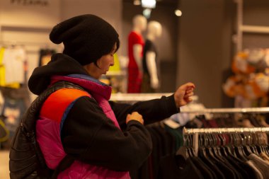 Alış veriş yapan kişi siyah süveteri inceliyor, parlak kabarık yelek ve bereyi görünür hale getiriyor, giyilmiş sırt çantası, sıcak mağaza ışığı, kumaşı hisseden eller, karar verirken samimi olma anı