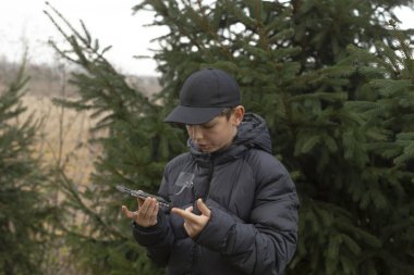 Kirpi ceketli çocuk dalları inceliyor, dikkatle bakıyor, elleri sopaları tutuyor, koyu şapka giyiyor, serin bulutlu arka plan, açık hava keşfi, düşünceli