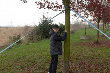 Ağaç destek kazıklarını denetleyen beyaz çocuk, mahalle parkında pratik gönüllü rolü, kalın ceket ve şapka giyiyor, genç ağaç ve ip desteklerinin yanında özenli duruş